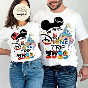 May include: White t-shirts with a Mickey Mouse ears design and the text "My First Disney Trip 2025". One shirt has a red bow and the other has a black bow.