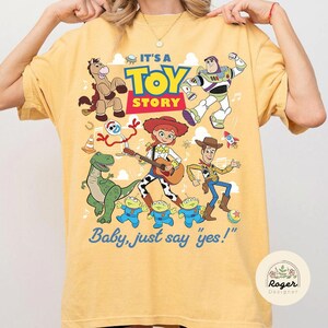 May include: A yellow short-sleeve t-shirt featuring a colorful graphic print of Toy Story characters including Woody, Jessie, Buzz Lightyear, Rex, Bullseye, Forky, and the aliens. The text reads IT'S A TOY STORY and Baby, just say 'yes!'