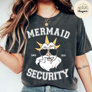 May include: Dark grey t-shirt with the words "MERMAID SECURITY" in white block letters. The shirt features a cartoon illustration of a bearded figure wearing sunglasses and a crown. The text "SINCE 2026" is also printed on the shirt.