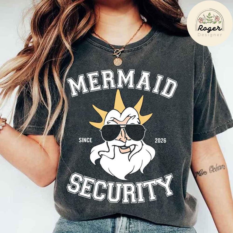 May include: Dark grey t-shirt with the words "MERMAID SECURITY" in white block letters. The shirt features a cartoon illustration of a bearded figure wearing sunglasses and a crown. The text "SINCE 2026" is also printed on the shirt.