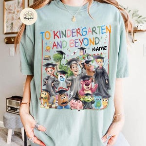 Peut inclure: Un t-shirt bleu clair avec le texte "TO KINDERGARTEN AND BEYOND" et le mot "NAME". Le t-shirt pr&eacute;sente un graphisme color&eacute; de personnages anim&eacute;s portant des chapeaux de remise de dipl&ocirc;mes. Les personnages incluent un dinosaure, un cow-boy et un ranger de l'espace.