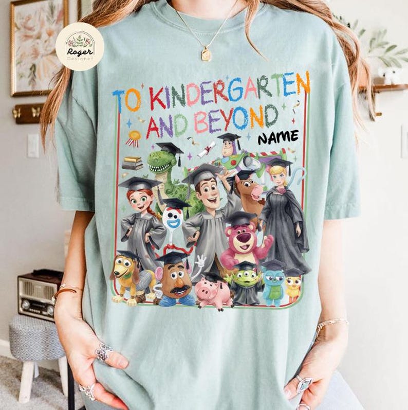 Peut inclure: Un t-shirt bleu clair avec le texte "TO KINDERGARTEN AND BEYOND" et le mot "NAME". Le t-shirt pr&eacute;sente un graphisme color&eacute; de personnages anim&eacute;s portant des chapeaux de remise de dipl&ocirc;mes. Les personnages incluent un dinosaure, un cow-boy et un ranger de l'espace.