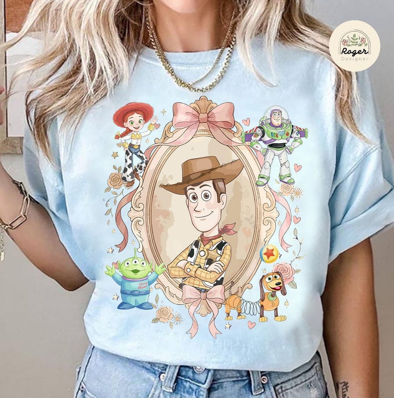 May include: Pale blue t-shirt with a cartoon illustration of Woody, Jessie, Buzz Lightyear, an alien, and Slinky Dog. The design is framed by a pink bow and decorative elements. The shirt has short sleeves.