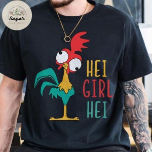 May include: Black t-shirt featuring a cartoon rooster with red, green, yellow, and white details. The text "HEI GIRL HEI" is printed in yellow, red, and teal. The shirt has a crew neck and short sleeves.