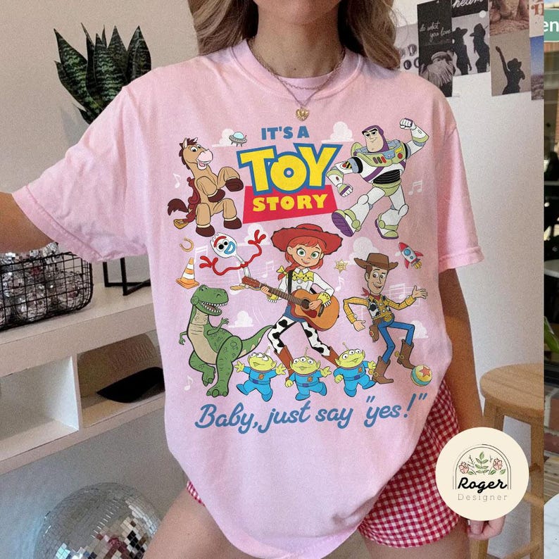 May include: A light pink t-shirt featuring a colorful graphic of Toy Story characters including Buzz Lightyear, Woody, Jessie, Rex, Bullseye, Forky, and the Aliens. The text reads IT'S A TOY STORY and Baby, just say 'yes!' in blue and yellow.