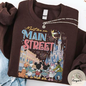 May include: A brown sweatshirt featuring a colorful graphic with the text "Meet me on Main Street." The design includes cartoon characters, a castle, fireworks, and a fairy. A silver necklace with a pendant is draped across the sweatshirt.