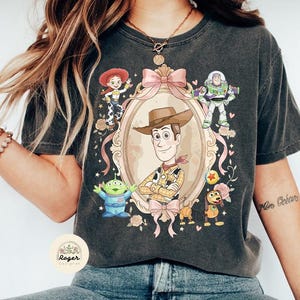 May include: Dark grey t-shirt with a framed illustration of Woody from Toy Story, surrounded by characters including Buzz Lightyear and Jessie. The design features pink ribbons and floral accents, giving a vintage look.