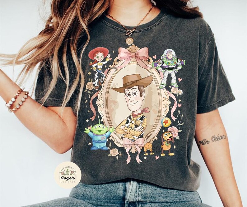 May include: Dark grey t-shirt with a framed illustration of Woody from Toy Story, surrounded by characters including Buzz Lightyear and Jessie. The design features pink ribbons and floral accents, giving a vintage look.