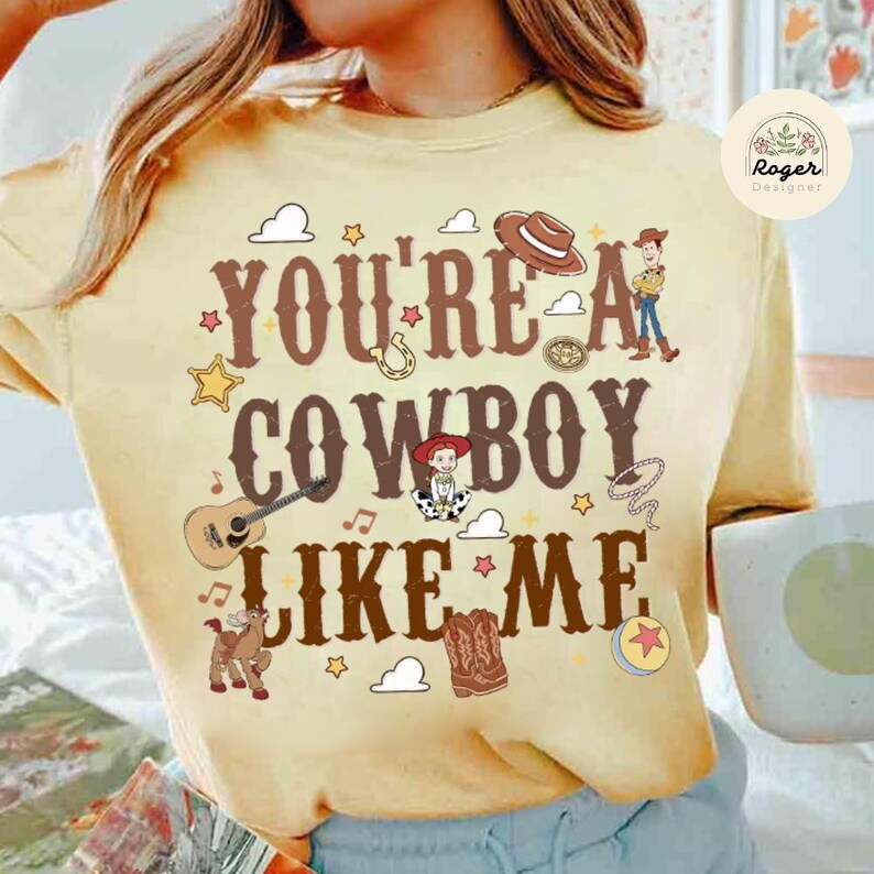 Może przedstawiać: Bladoz&oacute;łta bluza z okrągłym dekoltem z brązowym napisem "YOU'RE A COWBOY LIKE ME" otoczonym ilustracjami kowbojskiego kapelusza, gitary, lassa, konia, kowbojskich but&oacute;w, gwiazd, chmur i małej postaci z kresk&oacute;wki.