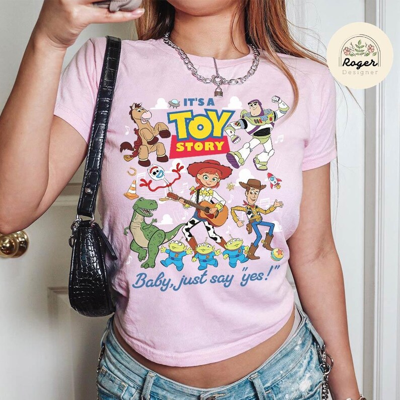 May include: A light pink t-shirt featuring a colorful graphic print of Toy Story characters including Woody, Buzz Lightyear, Jessie, Rex, Bullseye, and the Aliens. The text reads IT'S A TOY STORY and Baby, just say yes! with musical notes.