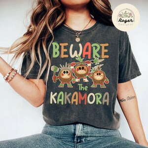 Puede incluir: Una camiseta gris oscuro con un estampado gr&aacute;fico de tres personajes de coco de dibujos animados con el texto "Beware The Kakamora".