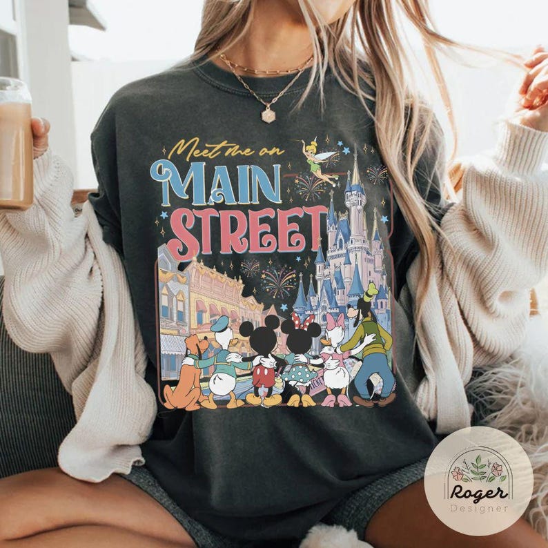 May include: Dark gray sweatshirt with a colorful graphic featuring Disney characters, a castle, fireworks, and the text "Meet me on Main Street." The design includes Mickey and Minnie Mouse, Donald Duck, and Goofy.