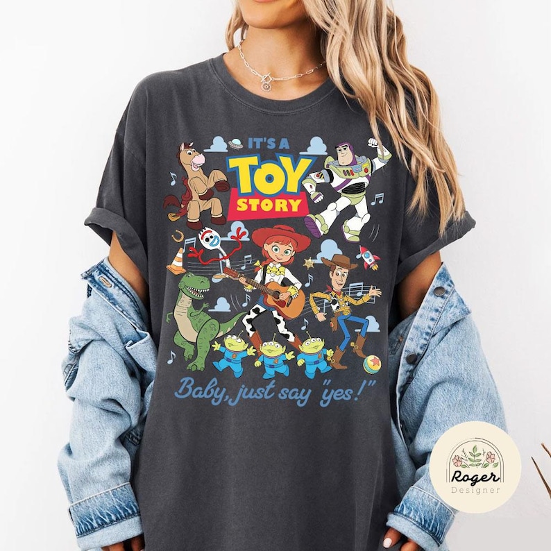 May include: A charcoal gray short-sleeve t-shirt featuring a colorful graphic print of Toy Story characters including Woody, Buzz Lightyear, Jessie, Rex, Bullseye, Forky, and the Aliens, with the text IT'S A TOY STORY and Baby, just say yes!