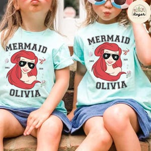 May include: Two light blue t-shirts with a mermaid graphic. The graphic features a red-haired mermaid wearing sunglasses, the word "MERMAID" above, and the name "OLIVIA" below. The shirts are worn by two young children.