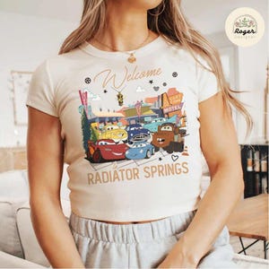 May include: Cream-colored t-shirt featuring a graphic of animated cars from the movie Cars, with the text "Welcome to Radiator Springs." The design includes a motel sign and various car characters.