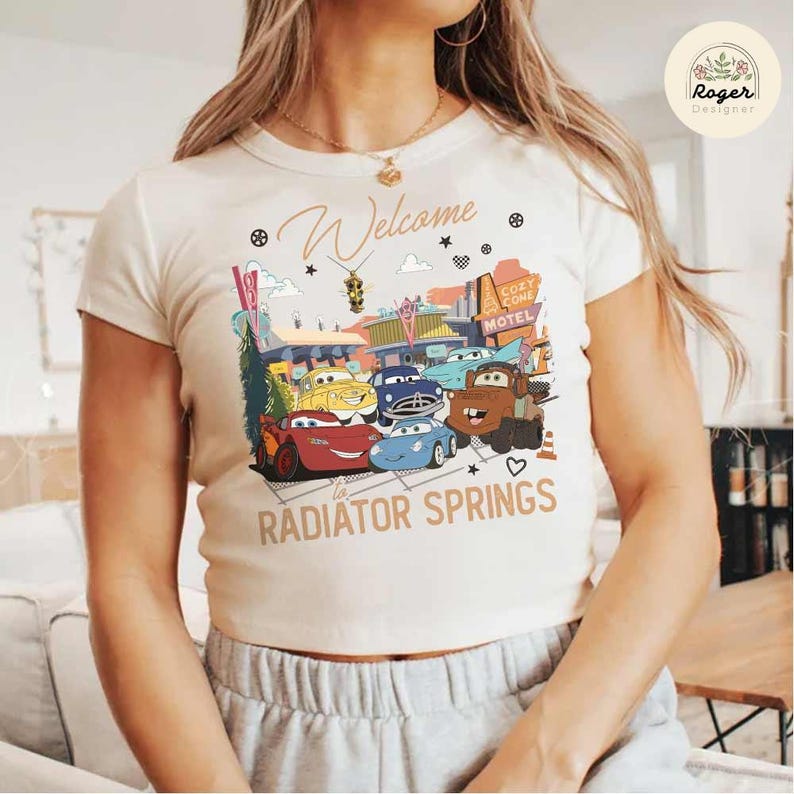 May include: Cream-colored t-shirt featuring a graphic of animated cars from the movie Cars, with the text "Welcome to Radiator Springs." The design includes a motel sign and various car characters.