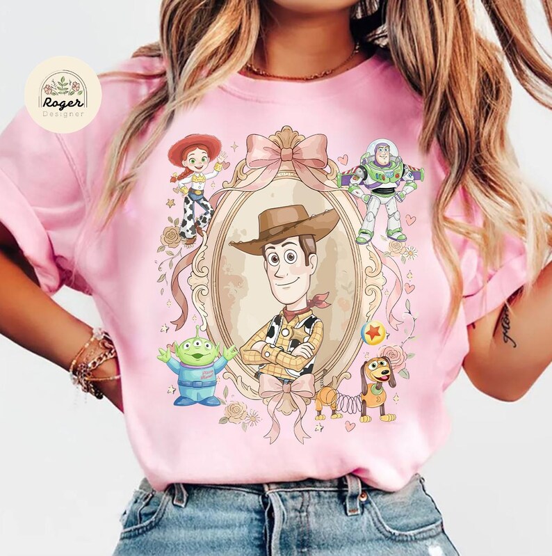 May include: Pale pink t-shirt with a central framed image of Woody from Toy Story, surrounded by characters including Buzz Lightyear, Jessie, and an alien. Floral details and a large bow complete the design.