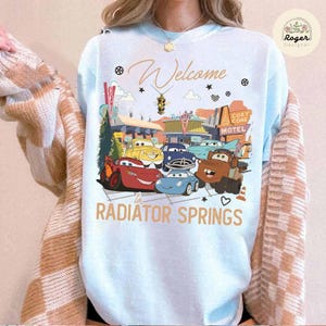 May include: Light blue sweatshirt featuring a colorful graphic of animated cars in front of a motel. The text "Welcome to Radiator Springs" is displayed in gold lettering. A cozy and fun design.