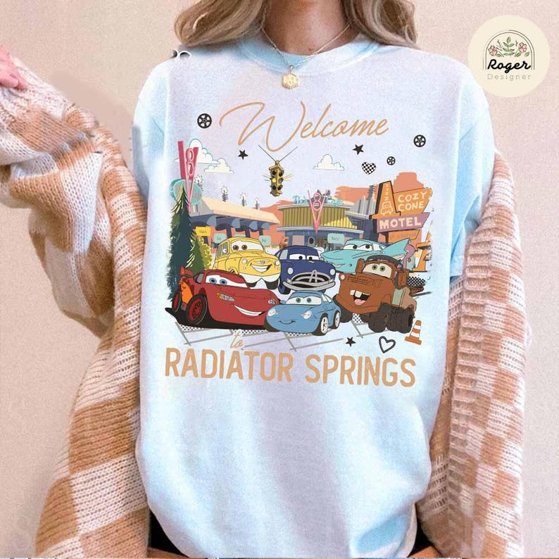 May include: Light blue sweatshirt featuring a colorful graphic of animated cars in front of a motel. The text "Welcome to Radiator Springs" is displayed in gold lettering. A cozy and fun design.