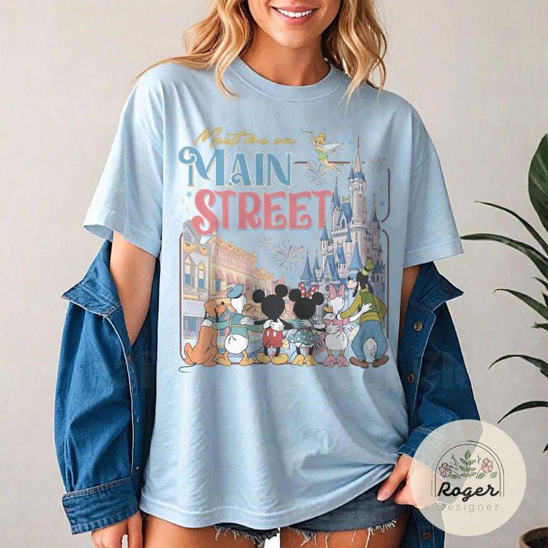 May include: Light blue t-shirt featuring a colorful graphic of Disney characters in front of a castle and the words "Meet me on Main Street". The design includes Mickey and Minnie Mouse, Donald Duck, and Goofy.