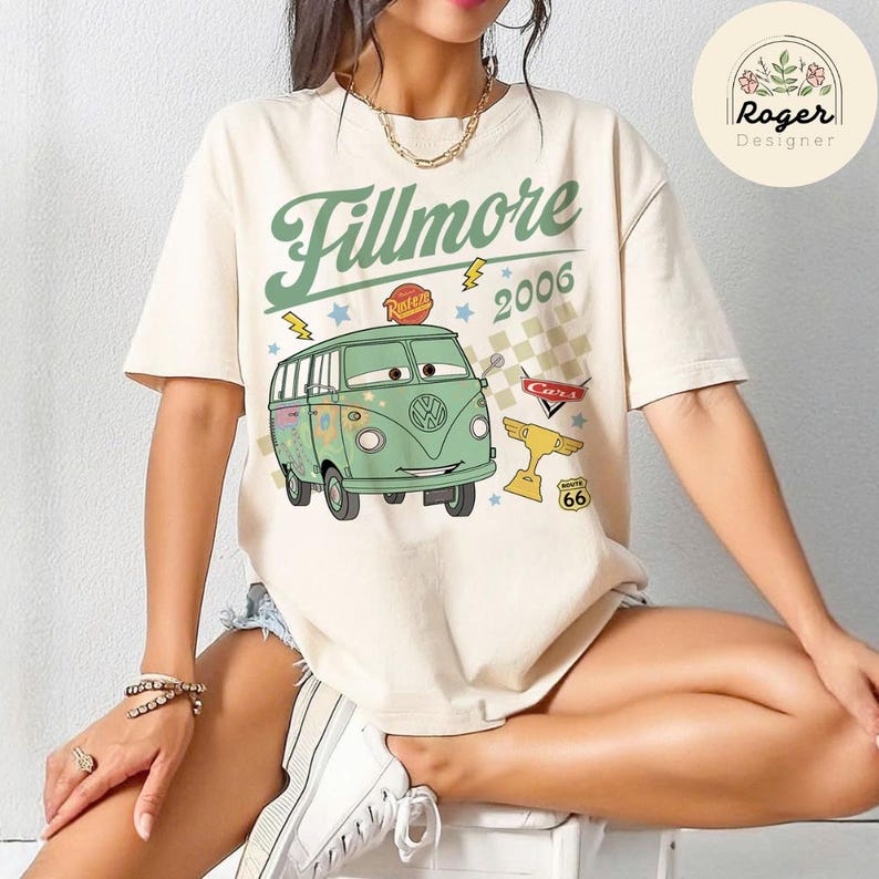 May include: Cream-colored t-shirt featuring a green cartoon van with the text "Fillmore 2006." The design includes lightning bolts, stars, and a Route 66 sign. The shirt has short sleeves and a crew neck.