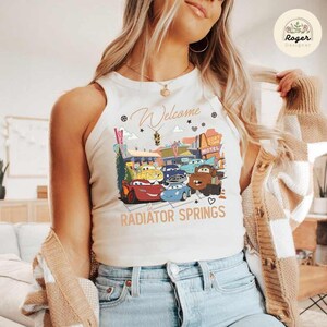 May include: Cream-colored tank top featuring a colorful graphic of characters from the movie Cars in front of a motel. The text "Welcome to Radiator Springs" is printed below the graphic.