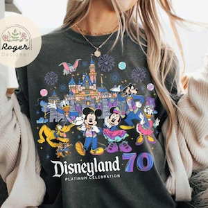 K&ouml;nnte beinhalten: Ein schwarzes T-Shirt mit einer bunten Grafik von Mickey Mouse und anderen Disney-Charakteren, die das 70-j&auml;hrige Jubil&auml;um von Disneyland feiern. Der Text "Disneyland Platinum Celebration" ist unter der Grafik gedruckt.