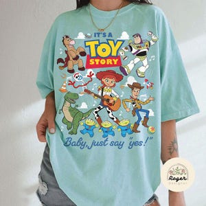 May include: A light mint green t-shirt featuring a colorful graphic print of Toy Story characters including Woody, Buzz Lightyear, Jessie, Bullseye, Rex, Forky, and the Aliens. The text reads IT'S A TOY STORY and Baby, just say yes!.