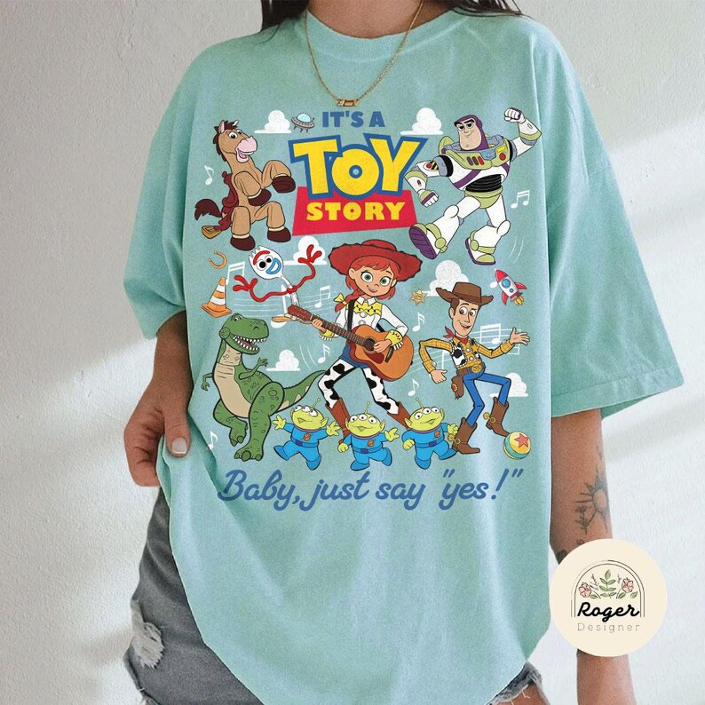 May include: A light mint green t-shirt featuring a colorful graphic print of Toy Story characters including Woody, Buzz Lightyear, Jessie, Bullseye, Rex, Forky, and the Aliens. The text reads IT'S A TOY STORY and Baby, just say yes!.
