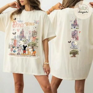 May include: An off-white t-shirt featuring a Walt Disney World graphic on the front with Mickey Mouse, Minnie Mouse, Donald Duck, and Pluto in front of the castle. The back displays a vertical arrangement of park icons including the castle.