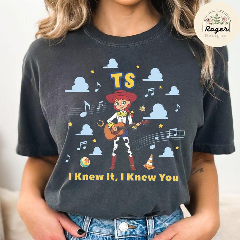 K&ouml;nnte beinhalten: Ein anthrazitfarbenes T-Shirt mit einer Grafik einer Cowgirl-Figur, die eine akustische Gitarre spielt. Das Design umfasst hellblaue Wolken, gelbe Sterne, Noten, einen Ball, einen Verkehrskegel sowie den Text TS und I Knew It, I Knew You.