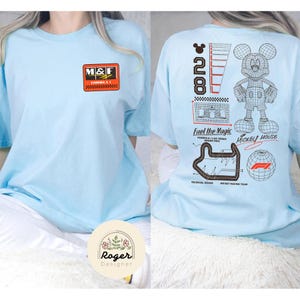 May include: Light blue t-shirt featuring a graphic design on the front and back. The front has a rectangular logo with the letters "M&F". The back features a Mickey Mouse graphic, the text "Fuel the Magic", and other racing-themed graphics.