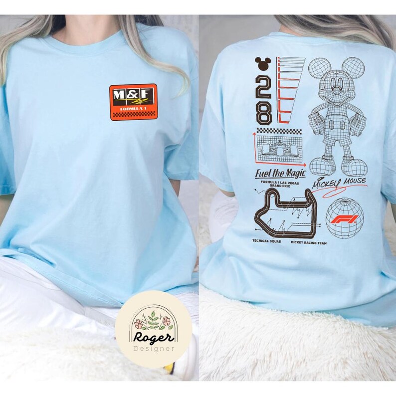 May include: Light blue t-shirt featuring a graphic design on the front and back. The front has a rectangular logo with the letters "M&F". The back features a Mickey Mouse graphic, the text "Fuel the Magic", and other racing-themed graphics.