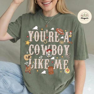 Może przedstawiać: Oliwkowozielony t-shirt z kr&oacute;tkim rękawem z napisem "YOU'RE A COWBOY LIKE ME" czcionką w stylu western. Wz&oacute;r zawiera ilustracje kapelusza kowbojskiego, gitary, konia, but&oacute;w, piłki oraz postaci z serii Toy Story na nadruku graficznym.
