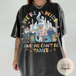 May include: Dark gray t-shirt with a colorful graphic featuring cartoon animal characters and the text "WE'RE WILD AND WE CAN'T BE TAMED." The design includes a cityscape and paw prints.