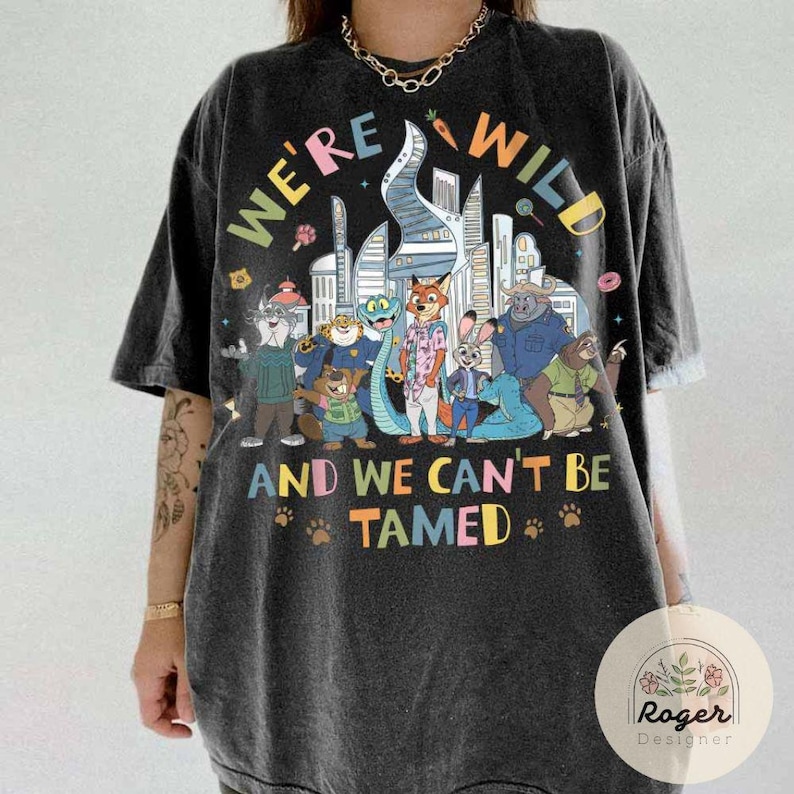 May include: Dark gray t-shirt with a colorful graphic featuring cartoon animal characters and the text "WE'RE WILD AND WE CAN'T BE TAMED." The design includes a cityscape and paw prints.