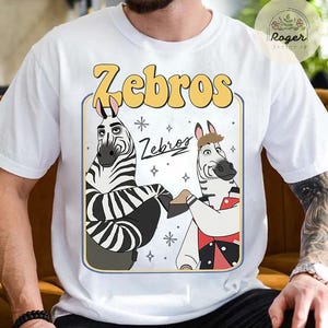 Pu&ograve; includere: T-shirt bianca con un design a cartoni animati di due zebre e la scritta "Zebros" in giallo. Le zebre sono in una cornice rettangolare con un bordo blu. La maglietta ha maniche corte.