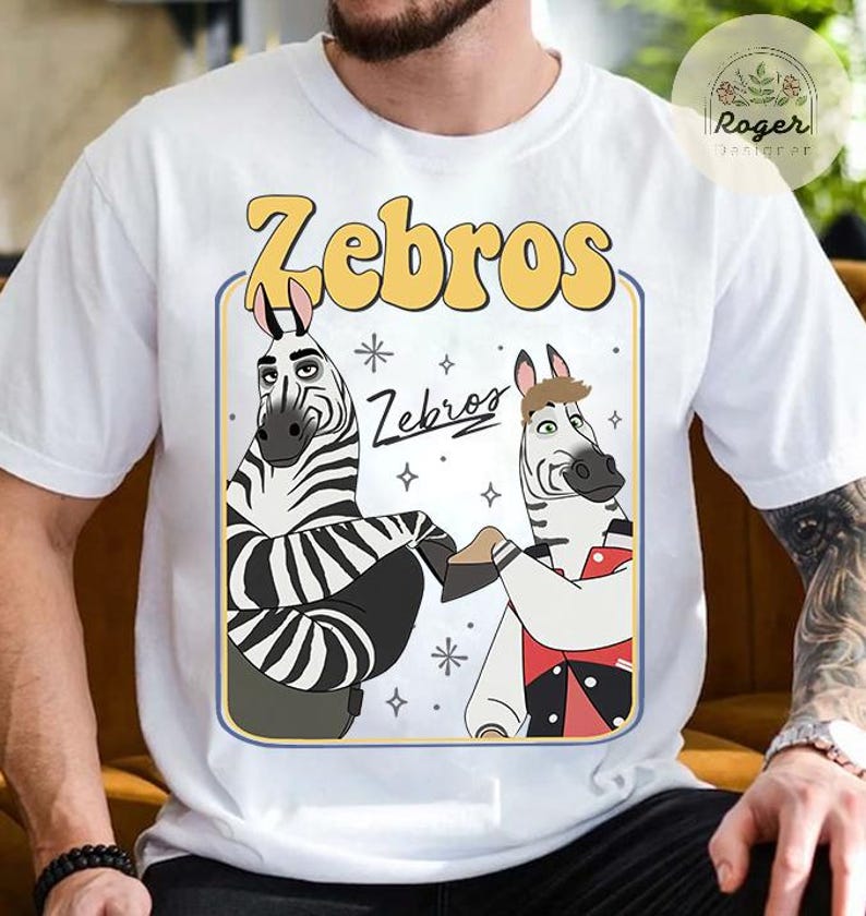 Pu&ograve; includere: T-shirt bianca con un design a cartoni animati di due zebre e la scritta "Zebros" in giallo. Le zebre sono in una cornice rettangolare con un bordo blu. La maglietta ha maniche corte.