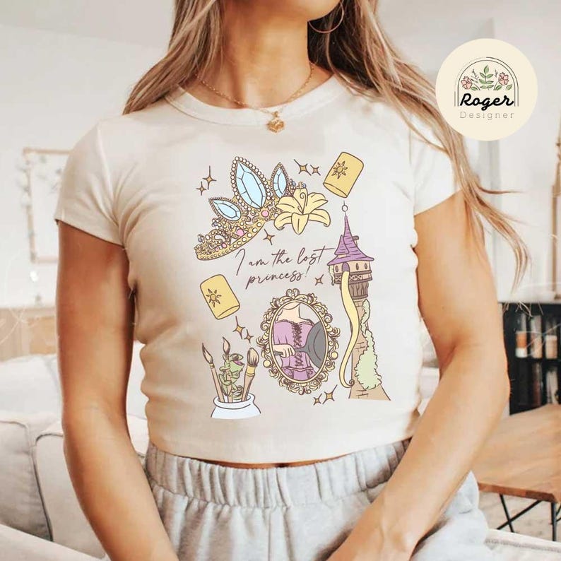 Może przedstawiać: Kremowy t-shirt z kolorowym nadrukiem graficznym i napisem "I am the lost princess!". Projekt zawiera tiarę, kwiat, wieżę i inne elementy. Koszulka jest z kr&oacute;tkim rękawem i okrągłym dekoltem.