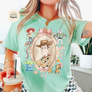 May include: A pale green t-shirt with a framed illustration of Woody from Toy Story, surrounded by characters including Buzz Lightyear, Jessie, and the Little Green Men. The design includes a pink bow and floral accents.