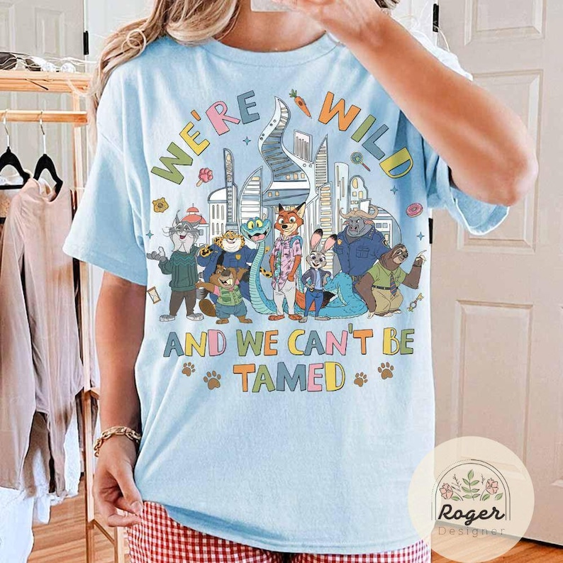 May include: Light blue t-shirt with the text "WE'RE WILD AND WE CAN'T BE TAMED" and a colorful graphic of cartoon animals in front of a city skyline. The shirt has short sleeves.