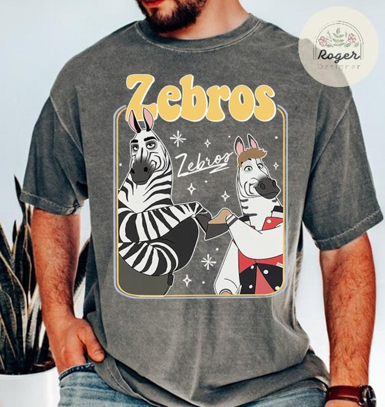 Pu&ograve; includere: T-shirt grigia con un disegno a cartoni animati di due zebre che si tengono per mano. La parola "Zebros" &egrave; in giallo sopra le zebre, con la stessa parola in un carattere pi&ugrave; piccolo sotto. Le zebre sono bianche e nere con orecchie rosa, in un rettangolo giallo.
