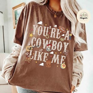 Może przedstawiać: Brązowy t-shirt z kr&oacute;tkim rękawem z napisem "YOU'RE A COWBOY LIKE ME" dużą, jasną czcionką. Wz&oacute;r zawiera ilustracje gitary, kowbojskiego kapelusza, gwiazdy szeryfa, buta, lassa oraz małej postaci z kresk&oacute;wki umieszczone na koszulce.