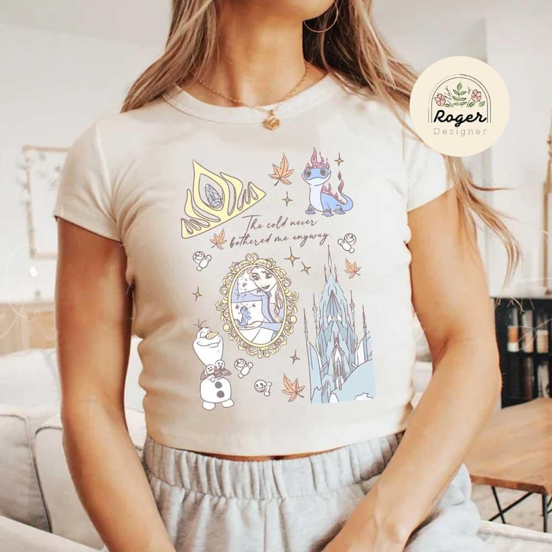Może przedstawiać: Kremowa koszulka z fantazyjną ilustracją z elementami z filmu Kraina Lodu. Projekt zawiera koronę, zamek, postacie i tekst "The cold never bothered me anyway."