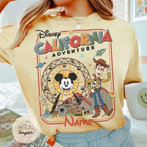 May include: Yellow t-shirt featuring a Disney California Adventure design. The graphic includes Mickey Mouse, Woody, a Ferris wheel, and the text "Name". The design is framed in a red border.