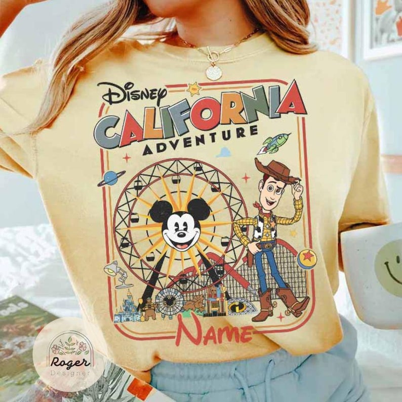May include: Yellow t-shirt featuring a Disney California Adventure design. The graphic includes Mickey Mouse, Woody, a Ferris wheel, and the text "Name". The design is framed in a red border.