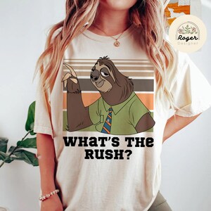 May include: Beige t-shirt featuring a cartoon sloth character with a green shirt and striped tie. The sloth is pointing with a smile, with the text "WHAT'S THE RUSH?" below. The design includes brown, orange, and green stripes.