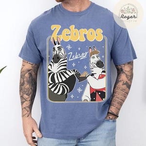 Pu&ograve; includere: T-shirt blu con un'illustrazione a cartoni animati di due zebre che si tengono per mano. La parola "Zebros" &egrave; in giallo sopra le zebre, con la parola "Zebros" in bianco sotto. Le zebre sono bianche e nere con caratteristiche da cartone animato.