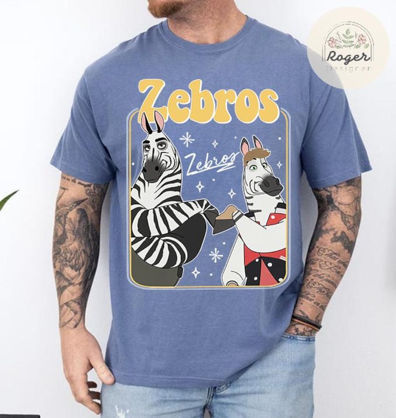 Pu&ograve; includere: T-shirt blu con un'illustrazione a cartoni animati di due zebre che si tengono per mano. La parola "Zebros" &egrave; in giallo sopra le zebre, con la parola "Zebros" in bianco sotto. Le zebre sono bianche e nere con caratteristiche da cartone animato.