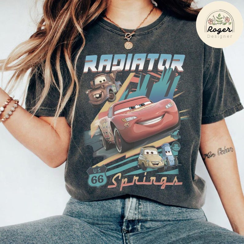 May include: A charcoal gray t-shirt featuring a graphic print of characters from the movie Cars, including Lightning McQueen and Mater. The design includes the text Radiator Springs and a US 66 road sign emblem, set against a stylized background.